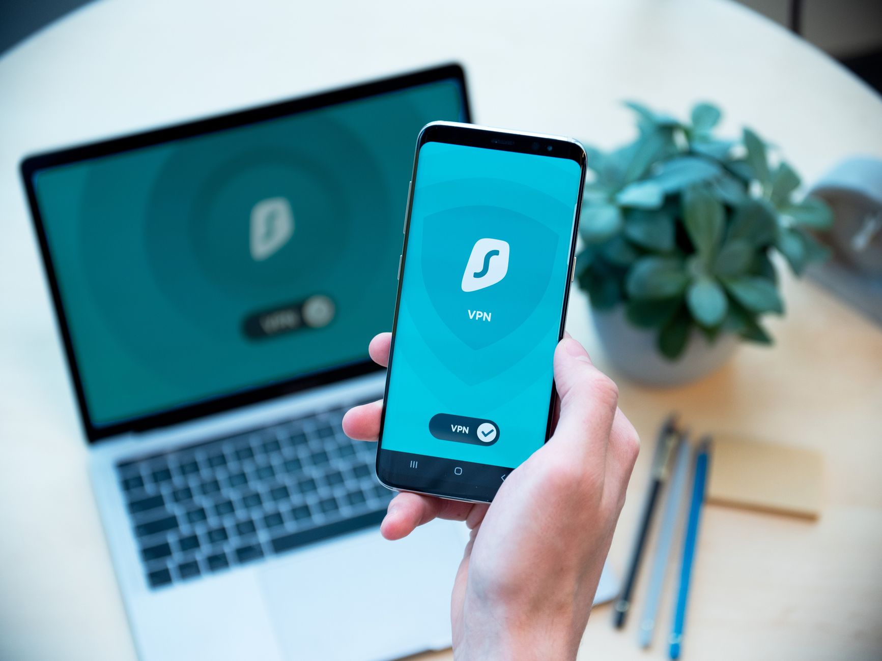 Elinde VPN uygulaması açık akıllı telefon tutan bir kişi ve arkada dizüstü bilgisayar